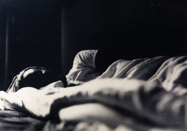 Black and white photograph of woman laying in in bed, partially hidden behind a blanket in the foreground.