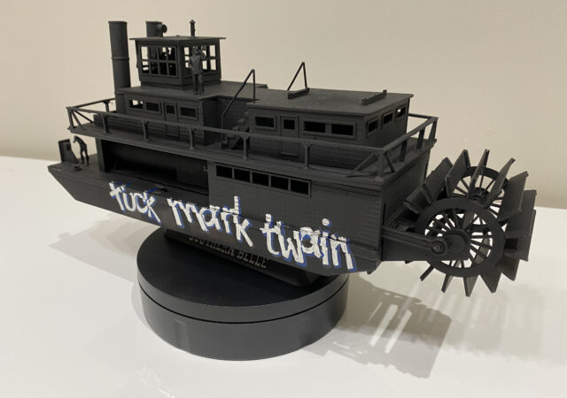 Sculpture of a miniature paddle boat painted black, with white text reading: 'fuck mark twain'