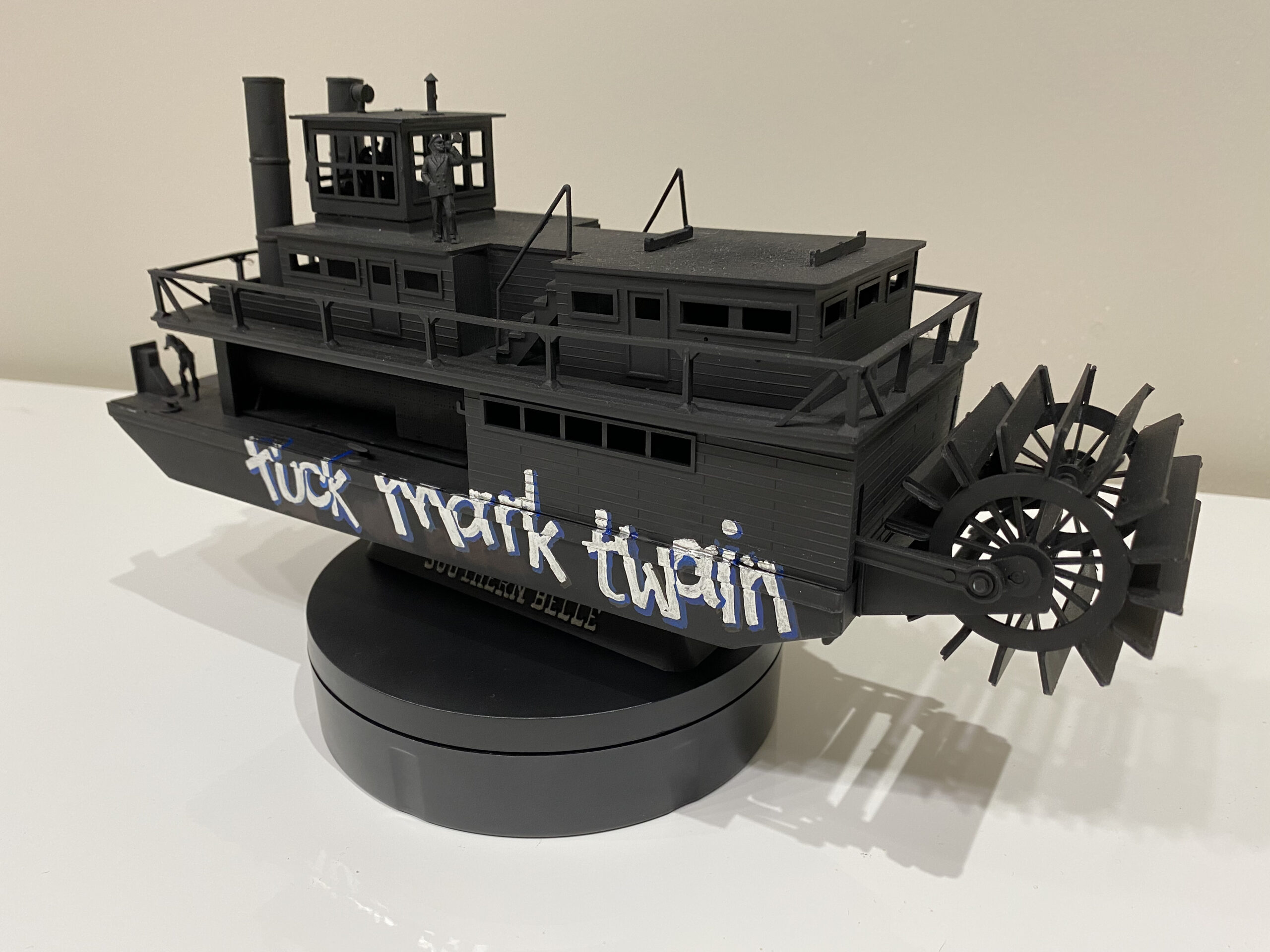 Sculpture of a miniature paddle boat painted black, with white text reading: 'fuck mark twain'